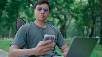 Businessman use smartphone and laptop sit on a chair in a park, Programmer hand hold smartphone conversation with abroad client distance meeting and typing on a laptop, Working online. video