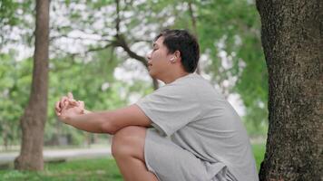 Zoom in Asian man sits on the ground outside crying and leaning against a tree. He is contemplating life, looking ahead with hope, and wearing EarPods while relaxing in a park. video