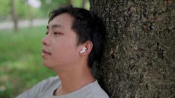 Close-up Asian man leaning on a tree thinking outside sitting on floor, He thinking about life, looking forward with hope and wearing AirPods while relaxing in a park, Healthy lifestyle. video