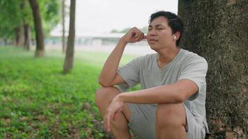Close-up Asian man sits on the ground outside, crying and leaning against a tree, thinking. He thinks about his life, looks forward with hope, and wears Air Pods while relaxing in a park. video