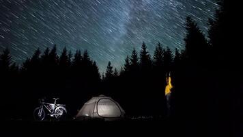 A man looks at the stars near the tent in the night forest. Star trail move in the night sky. Timelapse. Travel concept. 4K video