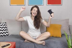 Brunet woman rejoices in the victory in the online game on the console. She is smile, sit relax on sofa at home have fun play online game. Close up photo