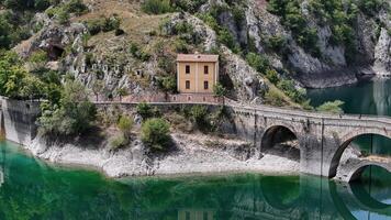 A quaint yellow house nestled between rocky hills and a serene emerald lake on a sunny afternoon photo