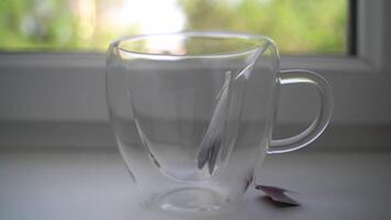 Person's hand puts tea bag in glass mug and pours boiling water over it, then dips it several times. Tea morning ceremony on windowsill in summer sunny day. video