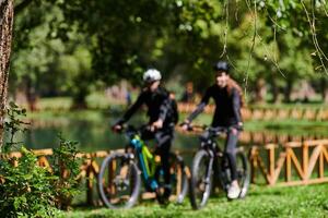 A blissful couple, adorned in professional cycling gear, enjoys a romantic bicycle ride through a park, surrounded by modern natural attractions, radiating love and happiness photo
