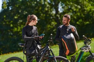 A sweet couple, equipped with bicycles and engrossed in coordinating their journey, checks their GPS mobile and watches while planning scenic routes in the park, seamlessly blending technology and photo
