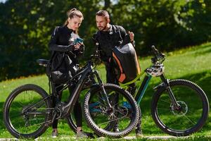 A sweet couple, equipped with bicycles and engrossed in coordinating their journey, checks their GPS mobile and watches while planning scenic routes in the park, seamlessly blending technology and photo