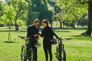 A sweet couple, equipped with bicycles and engrossed in coordinating their journey, checks their GPS mobile and watches while planning scenic routes in the park, seamlessly blending technology and photo
