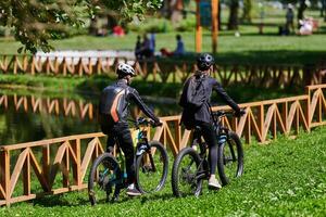A blissful couple, adorned in professional cycling gear, enjoys a romantic bicycle ride through a park, surrounded by modern natural attractions, radiating love and happiness photo