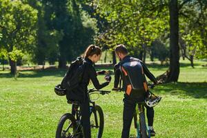 A blissful couple, adorned in professional cycling gear, enjoys a romantic bicycle ride through a park, surrounded by modern natural attractions, radiating love and happiness photo
