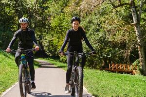 A blissful couple, adorned in professional cycling gear, enjoys a romantic bicycle ride through a park, surrounded by modern natural attractions, radiating love and happiness photo