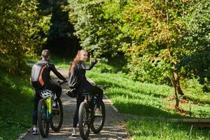 A blissful couple, adorned in professional cycling gear, enjoys a romantic bicycle ride through a park, surrounded by modern natural attractions, radiating love and happiness photo