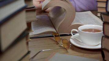 Female hand flipping through the pages of a book on table in library. Stacks of books, a cup of tea and glasses in the foreground. Concept of Reading and Education. Close up video