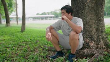 Asian man tired in airpods resting and breathing hard after working out at, He leaning against a tree and sitting outside on the ground thinking, Healthy Lifestyle. video