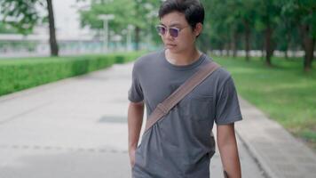 Asian man answers incoming phone call and walks talking on the phone in a public park. One hand holds a laptop and a bag. His legs are walking in nature to relax from work and study. video