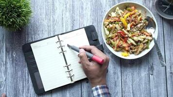 A man writing in a notebook with a pen on a table with a bowl of salad and a video