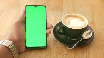A hand holding a smartphone with a green screen and a cup of coffee on a wooden table video