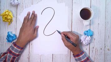 A man is writing the number 2 on a piece of paper with a pen and crumpled video
