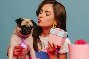 Beautiful young woman petting cute dog and holding gift box while sitting at the pink desk on blue background photo