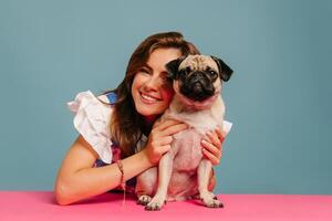 Happy young woman hugging cute pug dog while sitting at the pink desk on blue background photo