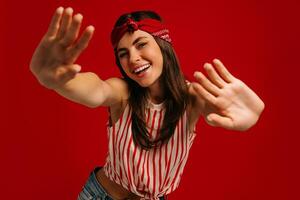 Joyful young hipster woman stretching out hands and smiling while standing on red background photo