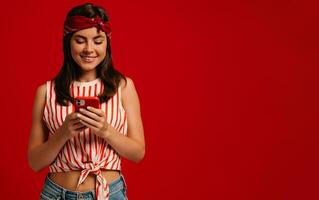 Stylish young hipster woman texting message on smart phone and smiling while standing on red background photo