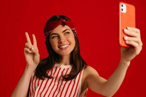 Joyful young hipster woman making selfie by mart phone and smiling while standing on red background photo