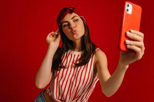 Playful young hipster woman making selfie by mart phone while standing on red background photo
