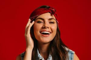 Cheerful young hipster woman with colorful make-up touching face and smiling on red background photo