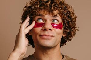 Handsome young curly man taking care of his skin while applying facial cosmetic mask on brown background photo