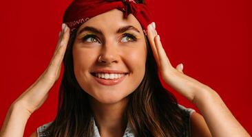 Attractive young hipster woman with colorful make-up touching face and smiling on red background photo
