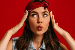 Beautiful young hipster woman with colorful make-up touching face and grimacing on red background photo