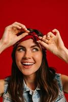 Attractive young hipster woman with colorful make-up adjusting bandana and smiling on red background photo