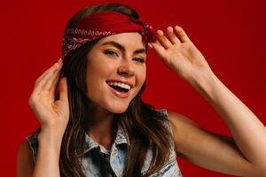 Attractive young hipster woman with colorful make-up adjusting bandana and smiling on red background photo
