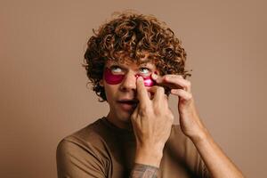 Handsome young curly man applying eye mask as facial treatment for better skin care on brown background photo