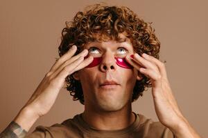 Handsome young curly man taking care of his skin while applying facial cosmetic mask on brown background photo