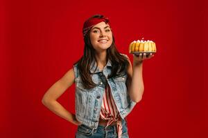 Happy young woman holding birthday cake and looking at camera with smile on red background photo
