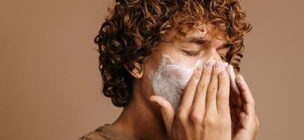 Handsome man enjoying beauty treatment while applying cream on face and standing on brown background photo