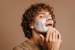 Handsome young man enjoying beauty treatment while applying cream on face and standing on brown background photo
