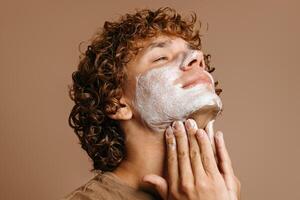 Handsome man enjoying beauty treatment while applying face cream and standing on brown background photo