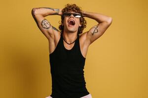 Excited young man in trendy glasses holding hands in hair and smiling against yellow background photo