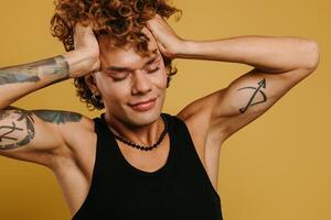 Young curly gay man with make-up holding hands in hair and keeping eyes closed against yellow background photo