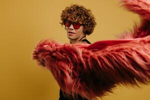 Curly young gay man in trendy glasses playing with fluffy coat against yellow background photo