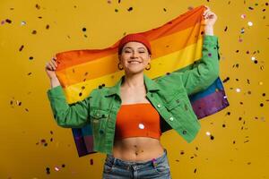 Happy woman carrying rainbow flag while confetti flying around her and against yellow background photo