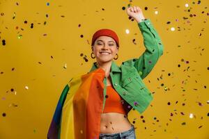 Happy woman carrying rainbow flag and gesturing while confetti flying against yellow background photo