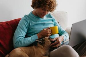 Young man sitting on the comfortable couch at home and petting dog while working from home photo