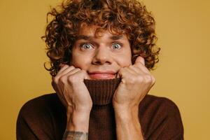 Playful curly young man stretching sweater neck and smiling at camera against yellow background photo