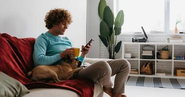 Handsome young curly man using smart phone while relaxing on the couch with his little dog together photo