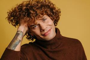 Handsome young curly man in sweater holding hand ion hair and smiling against yellow background photo