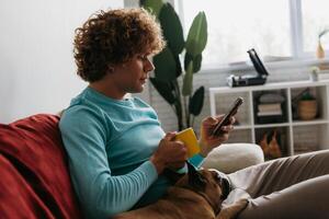 Handsome young curly man using smart phone while relaxing on the couch with his little dog together photo
